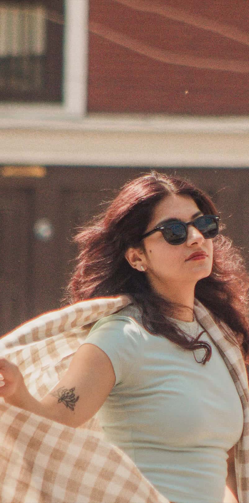 A woman in sunglasses opens a cream checked cashmere shawl on a street lined with red brick buildings and parked motorcycles
